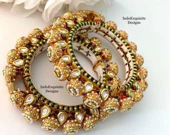 May include: Three gold bangles with intricate designs. The bangles are decorated with white pearls, green and red enamel, and clear stones. The bangles are stacked on top of each other.
