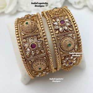 May include: Two gold bangles with intricate floral designs and red and green gemstones. The bangles are stacked on top of each other and have a beaded edge.