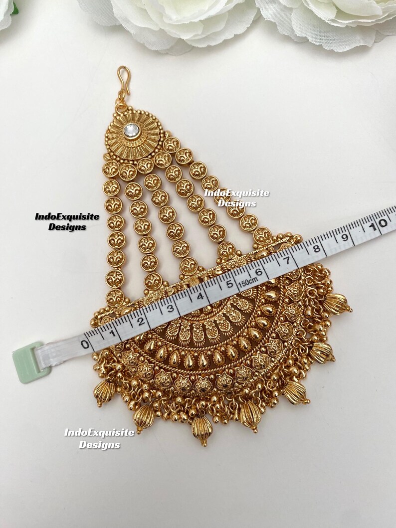 May include: A gold-colored headpiece with intricate floral and beaded designs. The headpiece is measured with a tape measure, showing a width of approximately 6 inches. The text "IndoExquisite Designs" is visible.