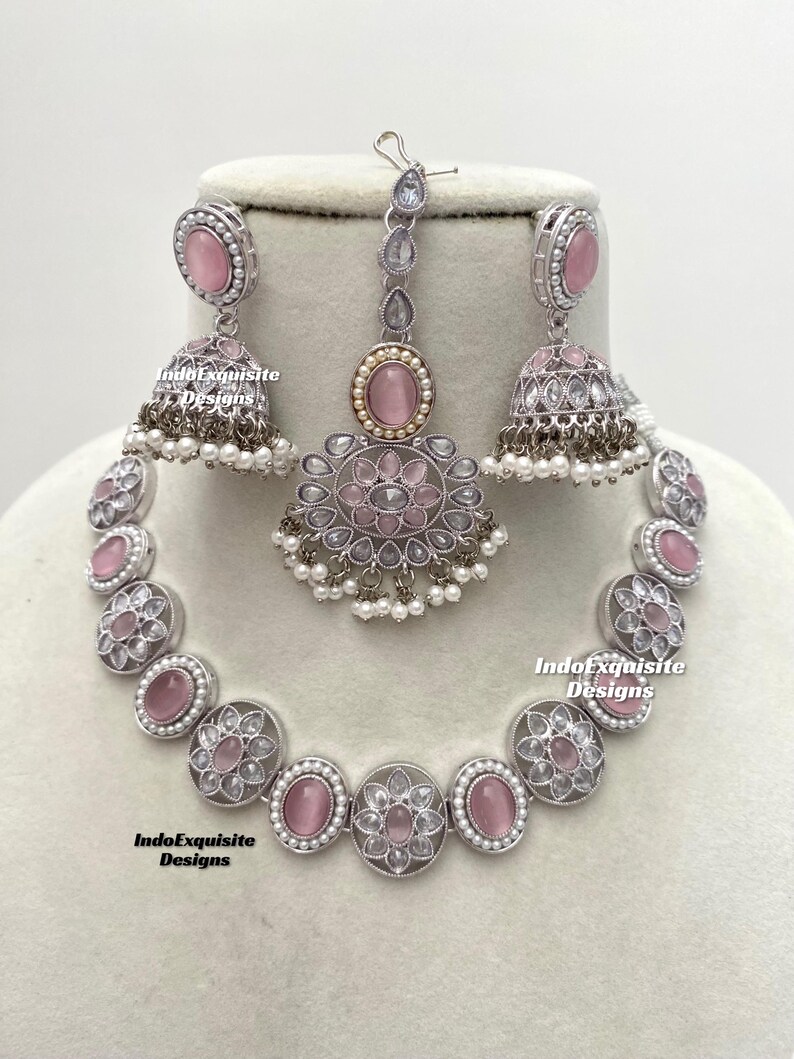 Op de afbeelding: Een zilveren en roze kettingset met een bijpassend paar oorbellen en een tikka. De ketting heeft een rij ronde roze stenen omgeven door zilveren filigraan en witte parels. De oorbellen en tikka hebben vergelijkbare ontwerpen.