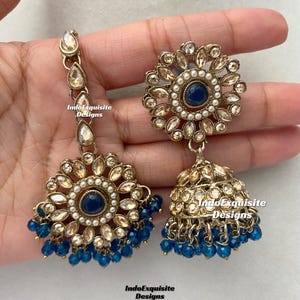May include: A pair of gold-tone earrings with blue and white accents. The earrings feature a round design with a blue center stone and a dangling jhumka with blue beads.