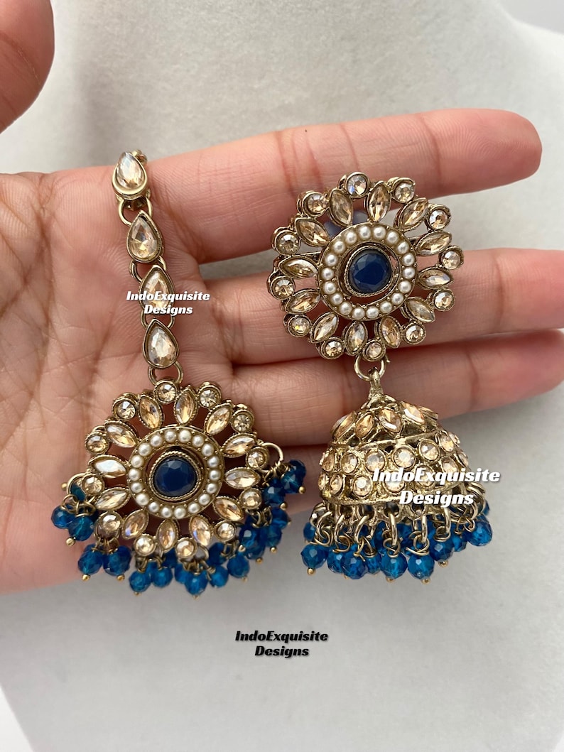 May include: A pair of gold-tone earrings with blue and white accents. The earrings feature a round design with a blue center stone and a dangling jhumka with blue beads.