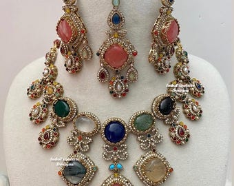 Puede incluir: Un conjunto de joyería que incluye un collar, dos pendientes y un maang tikka en tono dorado. Las piezas están adornadas con piedras multicolores rosas, azules, verdes y transparentes, rodeadas de pequeños cristales blancos en metal.
