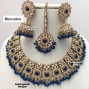 May include: A gold and blue Indian bridal necklace set with a large pendant, earrings, and a maang tikka. The necklace features a circular design with blue stones and pearls. The earrings are also circular with blue stones and pearls. The maang tikka is a small pendant with blue stones and pearls.