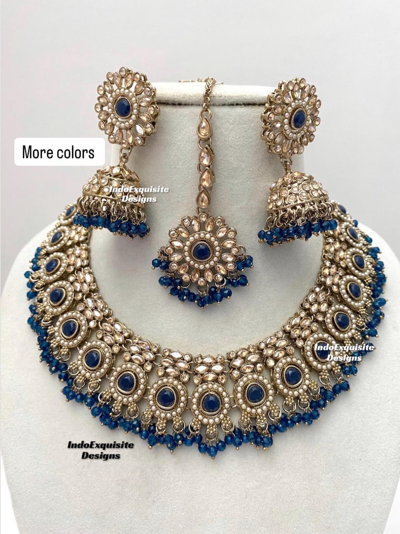May include: A gold and blue Indian bridal necklace set with a large pendant, earrings, and a maang tikka. The necklace features a circular design with blue stones and pearls. The earrings are also circular with blue stones and pearls. The maang tikka is a small pendant with blue stones and pearls.