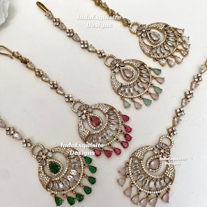 May include: Four gold-toned maang tikka headpieces featuring intricate metalwork, clear crystals, and teardrop-shaped stones in green, pink, and light pink. Each piece includes a hook for hair attachment and the text IndoExquisite Designs.