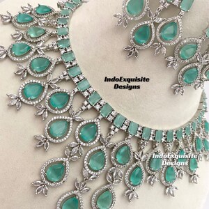 May include: A silver and green statement necklace and earrings set with teardrop-shaped stones. The necklace has a choker style with a row of rectangular stones and a cascading design of teardrop stones. The earrings are teardrop-shaped with a single stone.