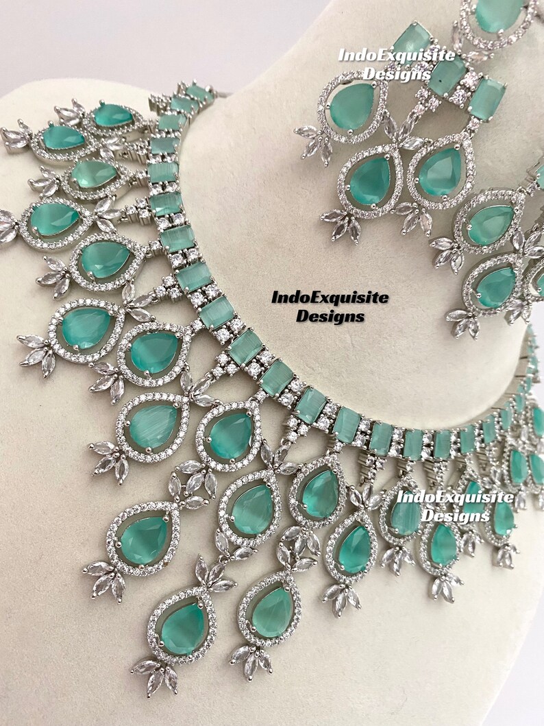 May include: A silver and green statement necklace and earrings set with teardrop-shaped stones. The necklace has a choker style with a row of rectangular stones and a cascading design of teardrop stones. The earrings are teardrop-shaped with a single stone.