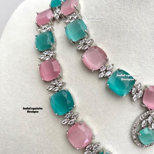 May include: A silver necklace with alternating pink and teal square-shaped stones. The stones are set in a silver frame with small clear stones around the edges. The necklace is on a white background. IndoExquisite Designs is written on the background.