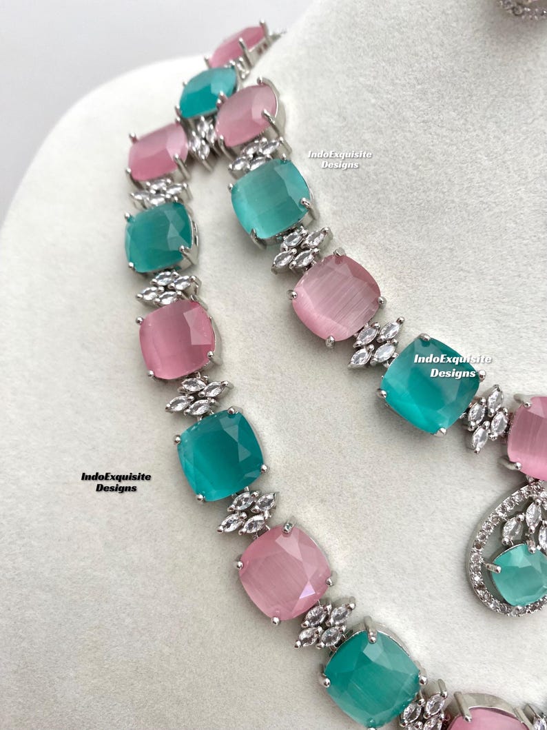 May include: A silver necklace with alternating pink and teal square-shaped stones. The stones are set in a silver frame with small clear stones around the edges. The necklace is on a white background. IndoExquisite Designs is written on the background.