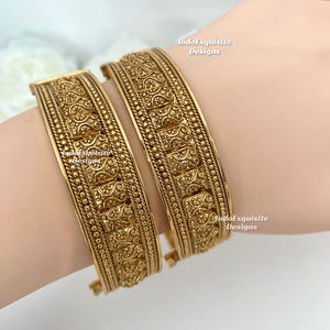 May include: Two gold bangles with detailed embossed patterns. The bangles are wide and feature intricate designs, including geometric shapes and beaded borders. The text "IndoExquisite Designs" is visible on the bangles.