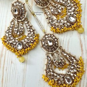 May include: A pair of gold earrings with yellow and white beads. The earrings are in the shape of a crescent moon and have a large, round, clear stone in the center. The earrings are decorated with intricate details and are perfect for a special occasion.