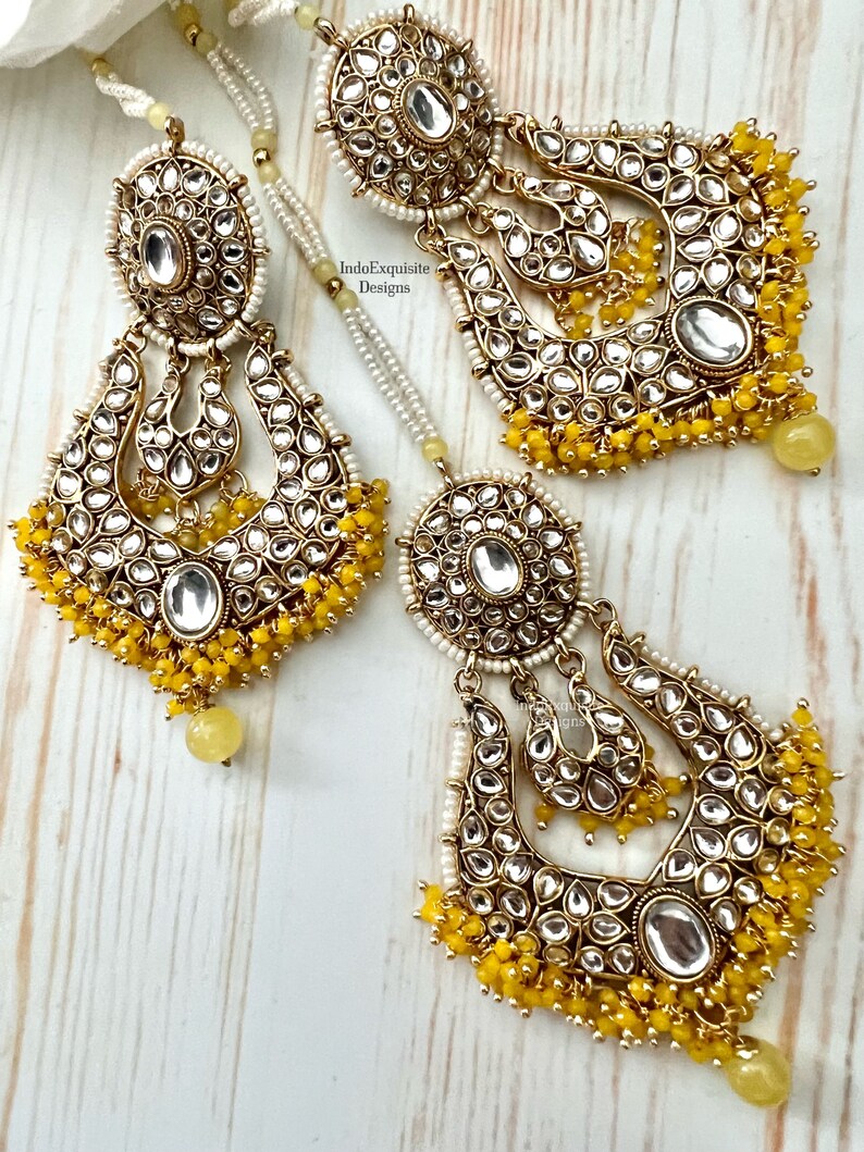 May include: A pair of gold earrings with yellow and white beads. The earrings are in the shape of a crescent moon and have a large, round, clear stone in the center. The earrings are decorated with intricate details and are perfect for a special occasion.