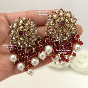 May include: Gold-coloured earrings with a floral design, featuring pink and red gemstones, small white pearls, and dangling red beads. The earrings are from IndoExquisite Designs.