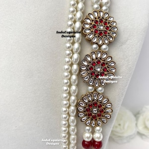 May include: A multi-strand necklace featuring white faux pearls and red beads. The necklace is adorned with three gold-toned floral pendants with red and clear crystal accents. The IndoExquisite Designs logo is visible.