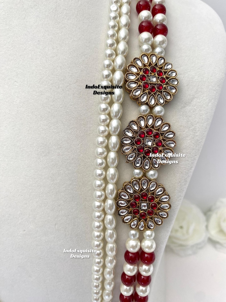 May include: A multi-strand necklace featuring white faux pearls and red beads. The necklace is adorned with three gold-toned floral pendants with red and clear crystal accents. The IndoExquisite Designs logo is visible.