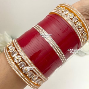 Può includere: Braccialetti rossi e dorati con pietre bianche e una fascia argentata. I braccialetti sono decorati con una fila di pietre grandi e trasparenti. Il testo "IndoExquisite Designs" è visibile sui braccialetti.
