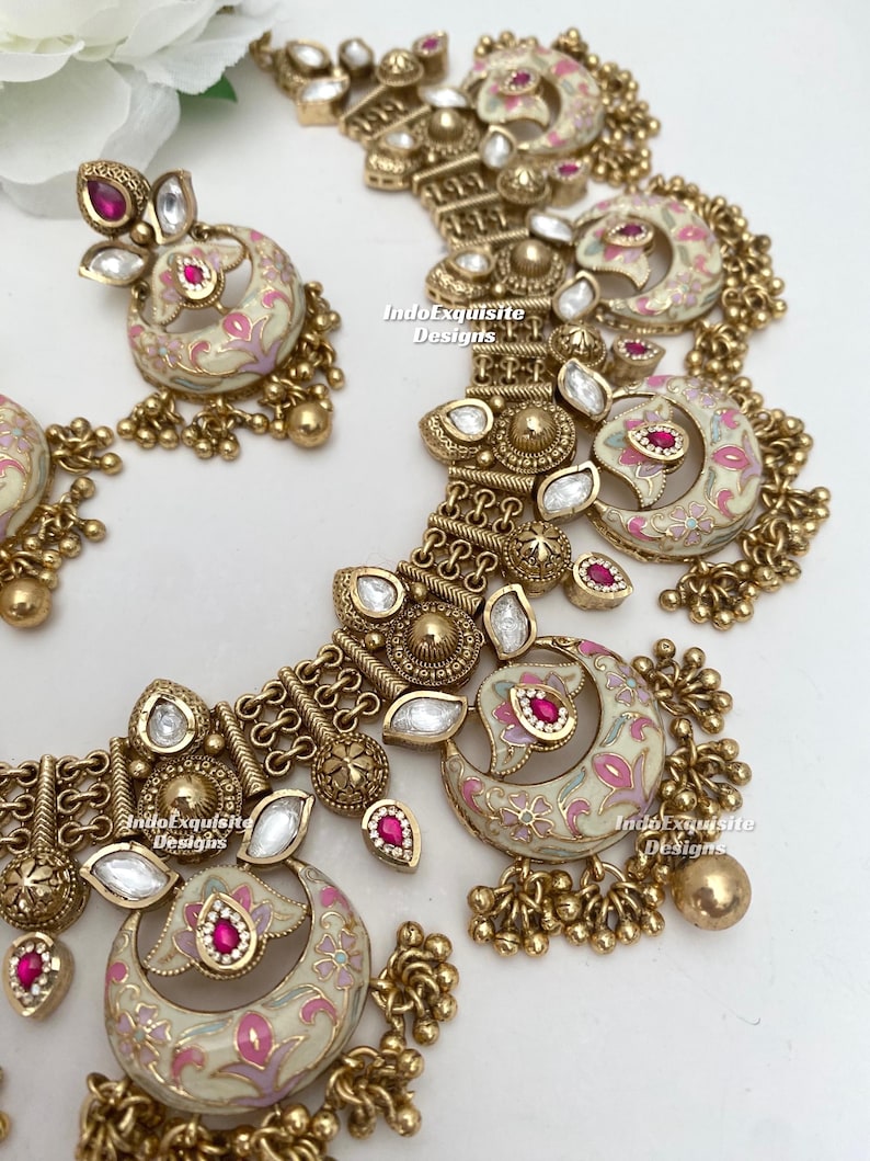 Pu&ograve; includere: Un set di collana e orecchini color oro. I gioielli presentano smalto color crema con disegni floreali rosa e accenti di pietre preziose rosse. Il set include perline dorate pendenti e pietre trasparenti a forma di goccia.