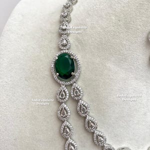 May include: A silver-toned necklace featuring a large, oval, emerald-green gemstone surrounded by a halo of small, clear crystals. The necklace has a delicate chain with teardrop-shaped crystal accents. The text "IndoExquisite Designs" is visible.