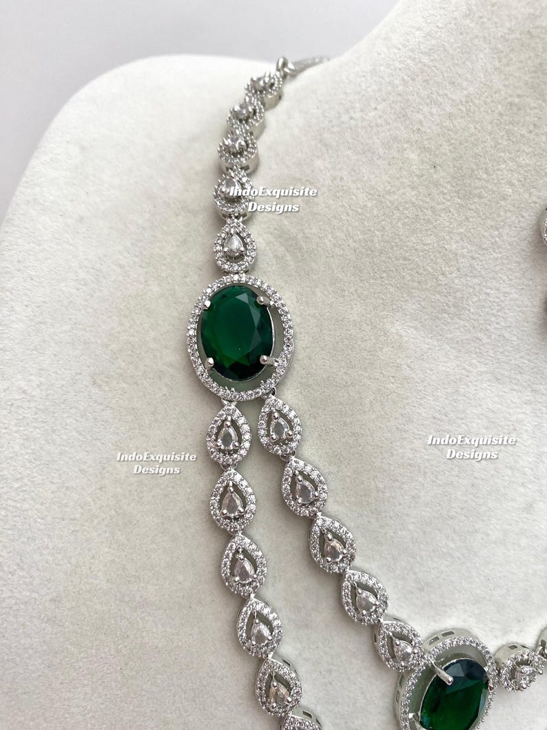 May include: A silver-toned necklace featuring a large, oval, emerald-green gemstone surrounded by a halo of small, clear crystals. The necklace has a delicate chain with teardrop-shaped crystal accents. The text "IndoExquisite Designs" is visible.