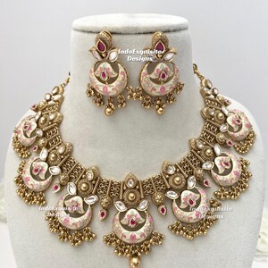 Pu&ograve; includere: Un set di collana e orecchini dorati con un motivo floreale color crema e rosa. Il set comprende una collana a pettorina e orecchini pendenti abbinati. Il testo "IndoExquisite Designs" &egrave; visibile.