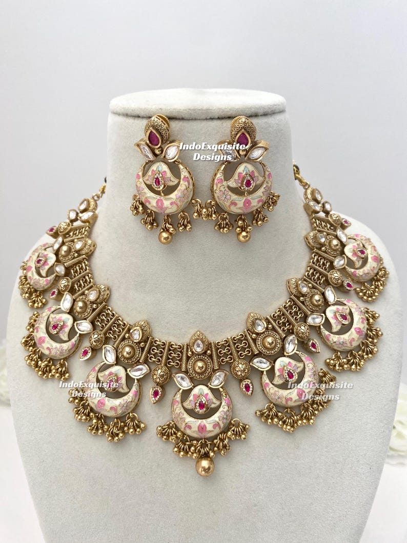 Pu&ograve; includere: Un set di collana e orecchini dorati con un motivo floreale color crema e rosa. Il set comprende una collana a pettorina e orecchini pendenti abbinati. Il testo "IndoExquisite Designs" &egrave; visibile.