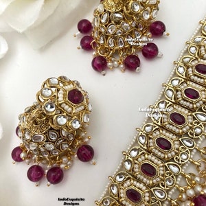 May include: A gold necklace with a geometric design and pink stones. The necklace is paired with matching earrings with a similar design. The earrings have a dangling design with pink stones.