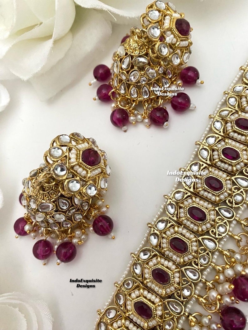 May include: A gold necklace with a geometric design and pink stones. The necklace is paired with matching earrings with a similar design. The earrings have a dangling design with pink stones.