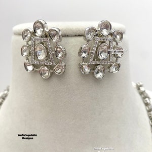 May include: A pair of silver earrings with a geometric design featuring clear stones. The earrings are on a white background.