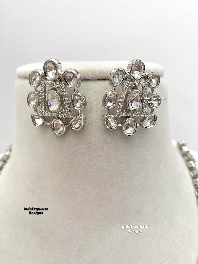May include: A pair of silver earrings with a geometric design featuring clear stones. The earrings are on a white background.