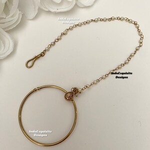 May include: Gold nose ring with a pearl chain and a teardrop-shaped pendant with a clear stone. The nose ring is open and has a hinged clasp. The chain is attached to the ring and has a small loop at the end. The pendant is attached to the chain and has a small pearl at the top.