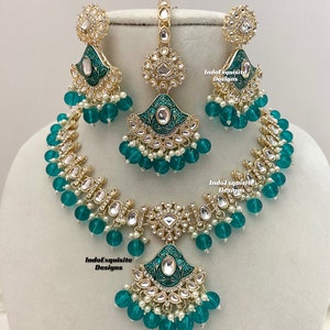 May include: A gold and green Indian bridal necklace set with a matching pair of earrings and a maang tikka. The necklace features a large pendant with a green enamel design and is adorned with pearls and clear stones. The earrings and maang tikka have similar designs.