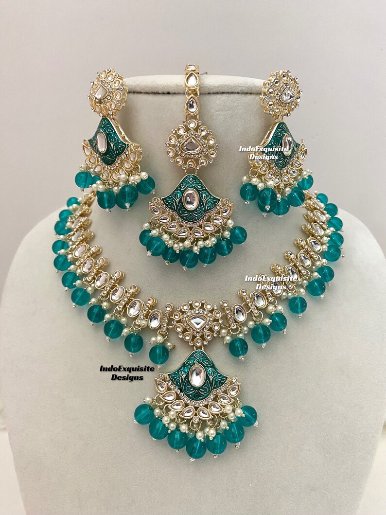 May include: A gold and green Indian bridal necklace set with a matching pair of earrings and a maang tikka. The necklace features a large pendant with a green enamel design and is adorned with pearls and clear stones. The earrings and maang tikka have similar designs.