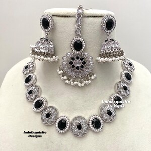 Op de afbeelding: Een zilveren ketting met zwarte en witte stenen, bijpassende oorbellen en een bijpassende maang tikka. De ketting heeft een cirkelvormig ontwerp met zwarte stenen in het midden van elke cirkel. De oorbellen zijn hangende oorbellen met een zwarte steen aan de onderkant. De maang tikka is een klein sieraad dat op het voorhoofd wordt gedragen. De sieraden zijn gemaakt van zilver en hebben een traditioneel Indiaas ontwerp.