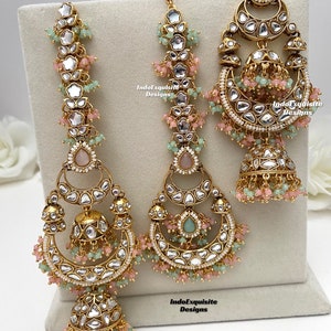 Puede incluir: Un conjunto de joyer&iacute;a en tono dorado que incluye dos pendientes largos y un maang tikka central. Cada pieza est&aacute; adornada con piedras claras, cuentas rosas y verde menta, y colgantes en forma de campana jhumka. El texto IndoExquisite Designs es visible.