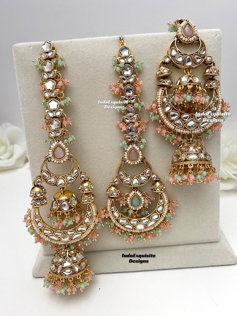 Puede incluir: Un conjunto de joyer&iacute;a en tono dorado que incluye dos pendientes largos y un maang tikka central. Cada pieza est&aacute; adornada con piedras claras, cuentas rosas y verde menta, y colgantes en forma de campana jhumka. El texto IndoExquisite Designs es visible.