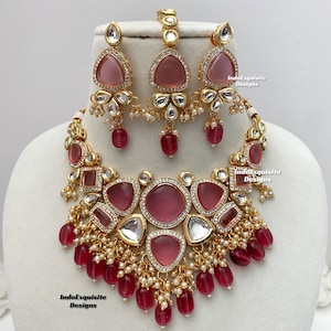 May include: An Indian bridal necklace set featuring a large pink stone pendant, smaller pink stones, and pearl accents. The set includes matching earrings and a tikka.