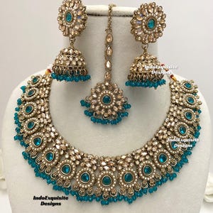 May include: A gold and teal Indian bridal necklace set with matching earrings and a tikka. The necklace features a large, round pendant with intricate details and a row of teal beads.