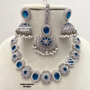 Op de afbeelding: Een zilveren ketting met blauwe en witte stenen, een bijpassend paar oorbellen en een bijpassende maang tikka. De ketting heeft een bloemmotief en is gemaakt van metaal.