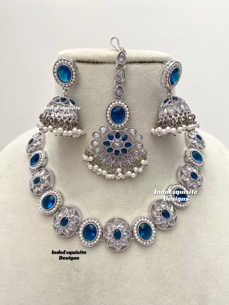 Op de afbeelding: Een zilveren ketting met blauwe en witte stenen, een bijpassend paar oorbellen en een bijpassende maang tikka. De ketting heeft een bloemmotief en is gemaakt van metaal.