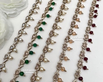 Puede incluir: Cinco cadenas decorativas metálicas con engastes en forma de lágrima, cristales transparentes y cuentas colgantes en blanco, verde, beige y rojo oscuro. Cada cadena muestra el texto IndoExquisite Designs impreso.