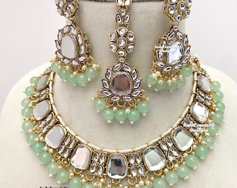 May include: A gold and silver necklace with green beads and mirror accents. The necklace is part of a jewelry set that also includes earrings and a maang tikka.