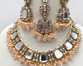 May include: A gold necklace with a white and gold design, featuring a row of large, mirrored, square-shaped stones. The necklace is accented with small, peach-colored beads. Matching earrings and a pendant are also included. The necklace is labeled "IndoExquisite Designs".