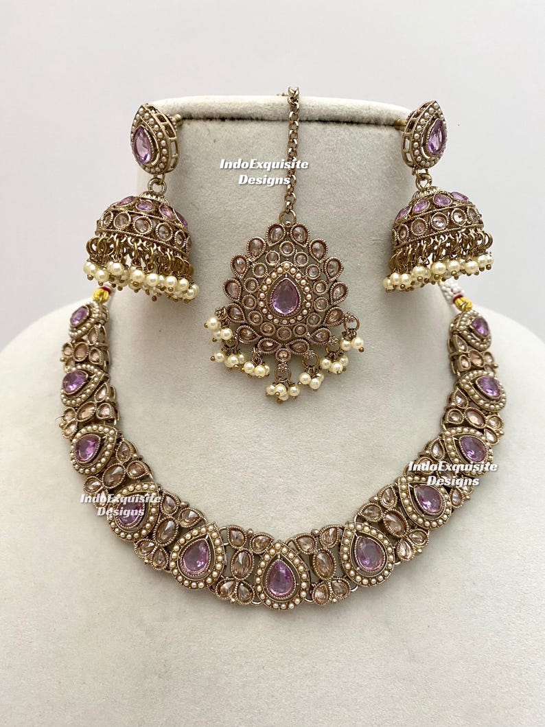 May include: A complete jewelry set featuring a necklace, earrings, and a headpiece. The set is antique gold-toned with lavender-colored stones and small white pearls. The earrings are bell-shaped, and the headpiece is a pendant.