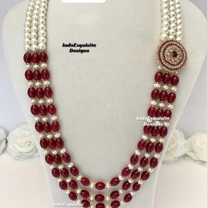May include: A multi-strand necklace with white pearls and oval, deep red beads. The necklace features a circular, ornate pendant with red and clear stones. The text "Design 3" and "IndoExquisite Designs" are visible.