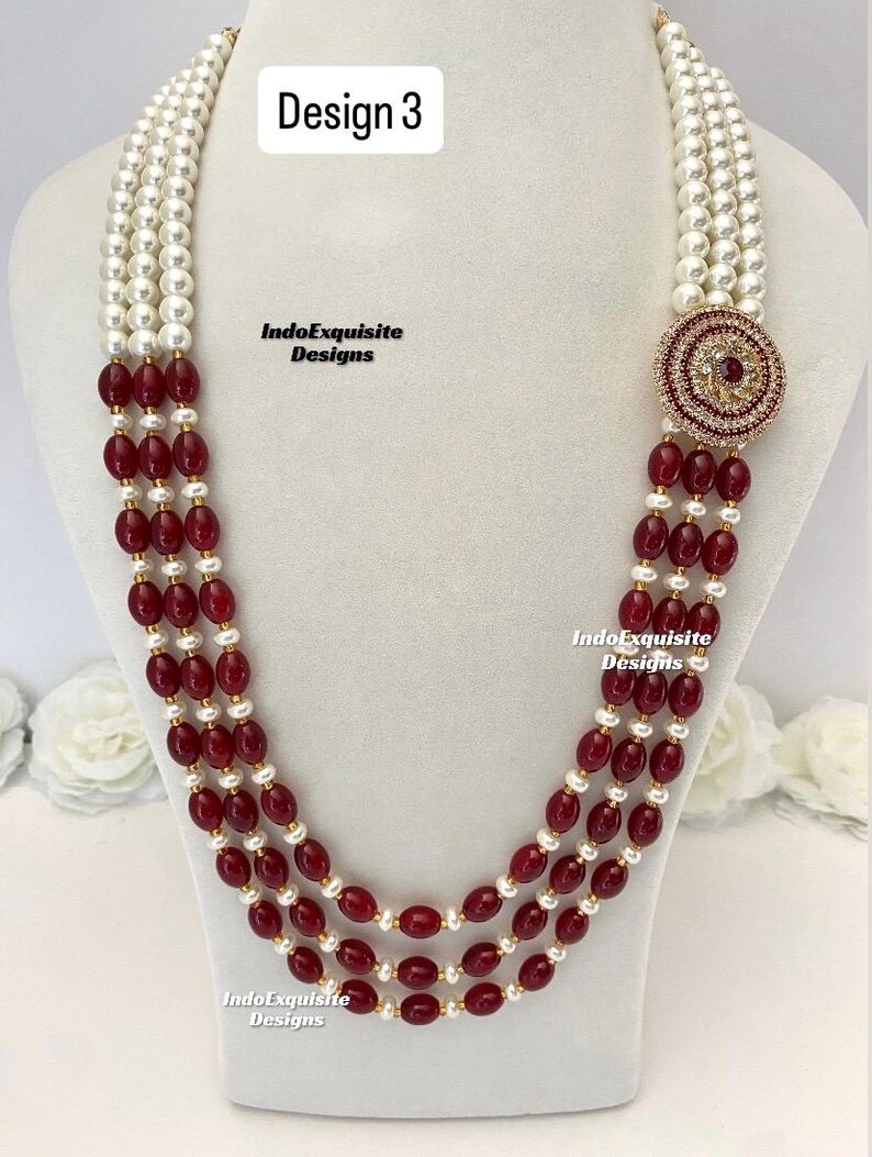 May include: A multi-strand necklace with white pearls and oval, deep red beads. The necklace features a circular, ornate pendant with red and clear stones. The text "Design 3" and "IndoExquisite Designs" are visible.