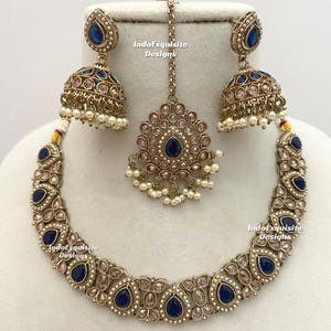 May include: A complete antique gold-toned jewelry set featuring a necklace, earrings, and a headpiece. The set includes teardrop-shaped navy blue gemstones and small white pearls. The earrings are bell-shaped. The text "IndoExquisite Designs" is visible.