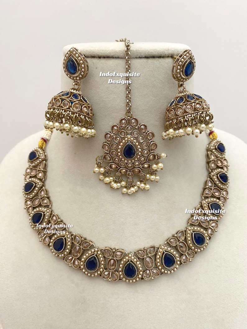 May include: A complete antique gold-toned jewelry set featuring a necklace, earrings, and a headpiece. The set includes teardrop-shaped navy blue gemstones and small white pearls. The earrings are bell-shaped. The text "IndoExquisite Designs" is visible.