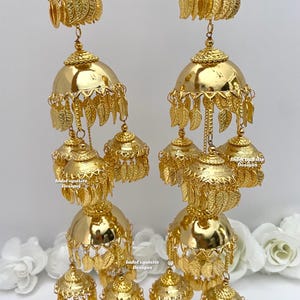 May include: A pair of gold-tone Indian bridal earrings with multiple tiers of dangling bells and leaf-shaped charms. The earrings are adorned with intricate details and have a traditional design.