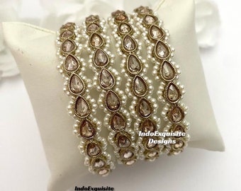 May include: Set of five gold bangles with clear and brown rhinestones and white pearl accents. The bangles are stacked on a white surface.